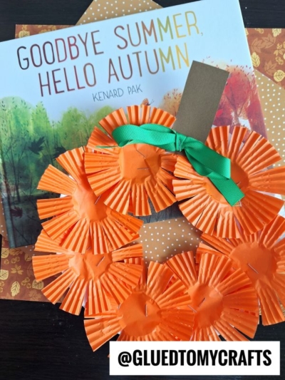 A children’s book titled Goodbye Summer, Hello Autumn lies next to a pumpkin craft made from orange cupcake liners, green ribbon, and brown paper—perfect for pairing with a festive pumpkin wreath—on patterned fall-themed paper. @GLUEDTOMYCRAFTS is printed at the bottom.