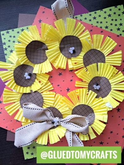 A charming cupcake liner sunflower wreath made from yellow cupcake liners, brown centers, and a beige ribbon, displayed on star-patterned paper. Username @GLUEDTOMYCRAFTS is in a green box at the bottom.
