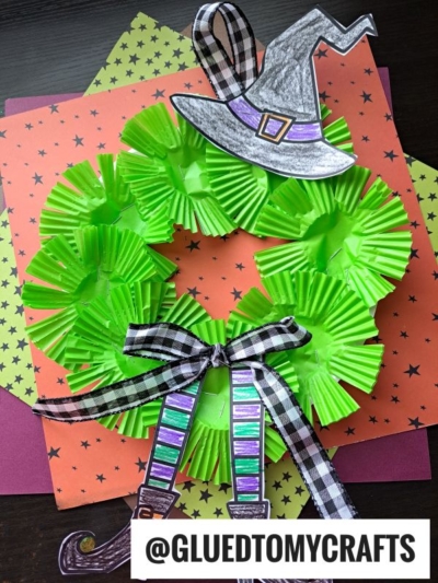 A witch wreath crafted from green cupcake liners, accented with a black-and-white plaid bow and whimsical paper witch hat and legs, displayed on star-patterned paper. Text: @GLUEDTOMYCRAFTS.