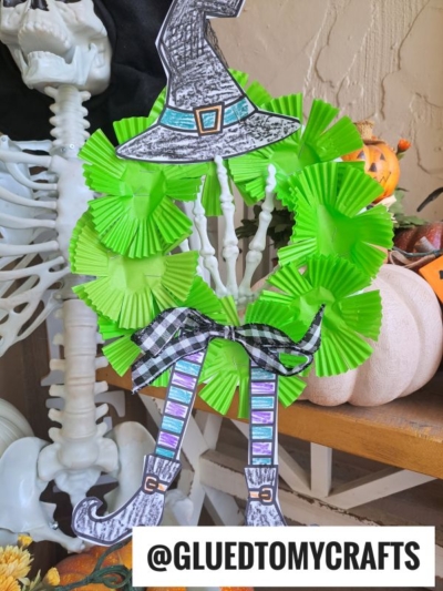 A skeleton holds a whimsical witch wreath made of bright green cupcake liners, adorned with a witch hat, striped legs, and a black plaid bow. Pumpkins and autumn decor complete the festive scene. Text reads @GLUEDTOMYCRAFTS.