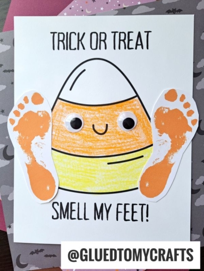 A footprint candy corn with googly eyes and orange-painted footprints for feet is on white paper. The text reads TRICK OR TREAT SMELL MY FEET! with the handle @GLUEDTOMYCRAFTS at the bottom.
