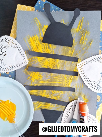 A bumblebee craft made from black paper shapes, yellow fork painting crafts, and paper doilies on a gray background. A plate with yellow paint, a glue stick, and paintbrush are nearby. @GLUEDTOMYCRAFTS is written at the bottom.