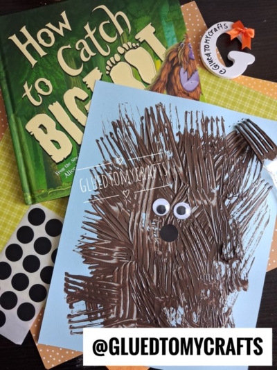 A children’s book, a fork-painted brown Bigfoot craft with googly eyes on blue paper, black dot stickers, and a Glued To My Crafts label are displayed on a yellow patterned background—highlighting the fun of fork painting crafts for kids.