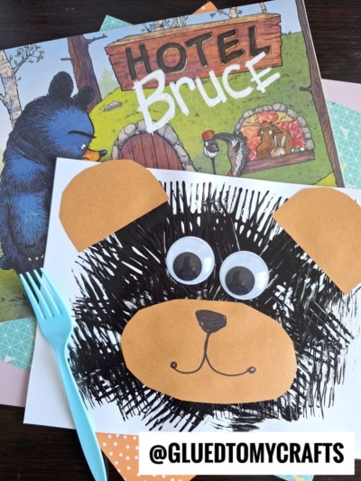 A bear craft made from brown paper, a white sheet, large googly eyes, and black paint is displayed next to the book Hotel Bruce. A blue plastic fork highlights this fun fork painting crafts idea with patterned paper. @GLUEDTOMYCRAFTS is written at the bottom.