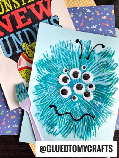 A blue monster craft, created with fork painting crafts techniques on light blue paper, features googly eyes and a drawn mouth. It sits beside a book and plastic fork. @GLUEDTOMYCRAFTS is written at the bottom.