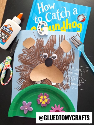 A homemade craft of a groundhog made from paper and a paper plate sits on a table with scissors, glue, and the book How to Catch a Groundhog. Foam flower stickers decorate the grass, perfect for adding to fork painting crafts fun.