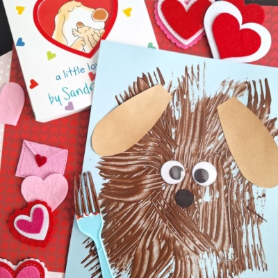 A children’s craft scene with a brown dog made from fork painting crafts, googly eyes, and paper ears on blue paper, a book titled “Snuggle Puppy,” felt hearts, a plastic fork, and a red heart-patterned background.