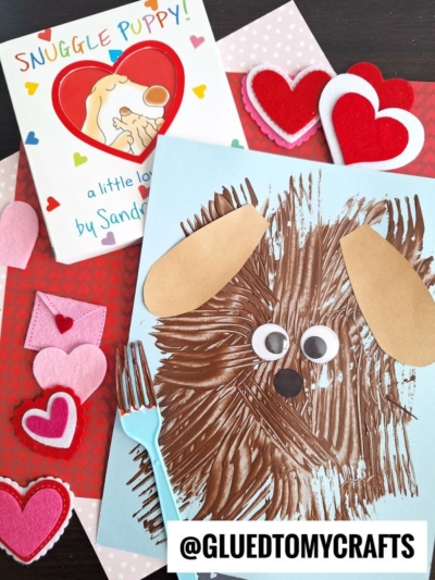 A children’s craft scene with a brown dog made from fork painting crafts, googly eyes, and paper ears on blue paper, a book titled “Snuggle Puppy,” felt hearts, a plastic fork, and a red heart-patterned background.
