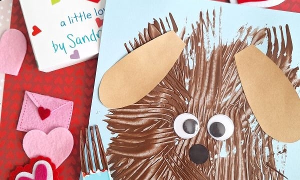 A children’s craft scene with a brown dog made from fork painting crafts, googly eyes, and paper ears on blue paper, a book titled “Snuggle Puppy,” felt hearts, a plastic fork, and a red heart-patterned background.