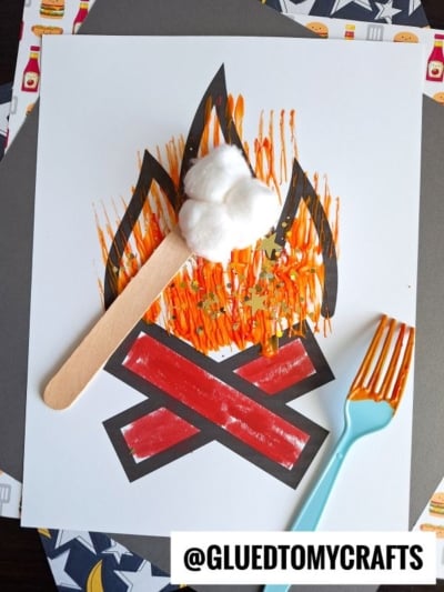 A paper campfire craft featuring a painted paper flame in orange, yellow, and brown strokes uses fork painting crafts techniques. Cotton ball marshmallows on a craft stick and a painted plastic fork complete the @GLUEDTOMYCRAFTS artwork.