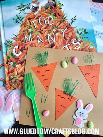 A children’s book titled Too Many Carrots sits behind a carrot-themed craft made with orange paper triangles, fork painting crafts for the green tops, small decorative eggs, and a bunny cutout, with a green plastic fork nearby.