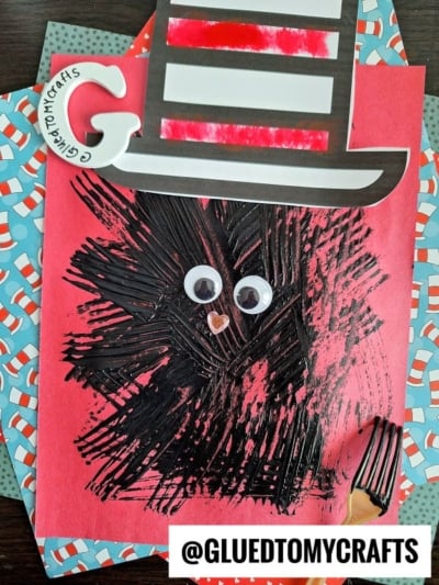 A children’s craft featuring the Cat in the Hat’s hat made of paper, with fork painting crafts creating a furry face, two googly eyes, and a small orange nose on red paper. A plastic fork and @GLUETOMYCRAFTS are also shown.