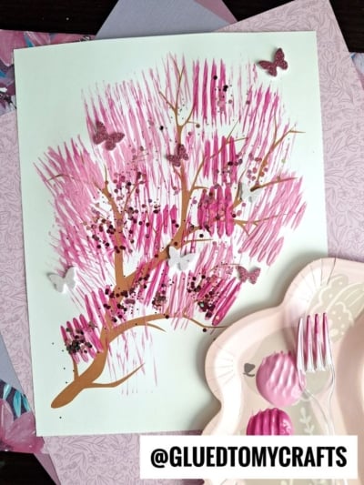 A handmade cherry blossom tree artwork showcases fork painting crafts with pink paint strokes for blossoms, brown branches, black dots for detail, and glittery butterfly stickers. Pink utensils and plates are placed nearby. The text @GLUEDTOMYCRAFTS appears at the bottom.