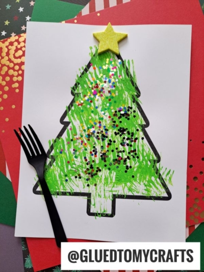 A Christmas tree fork painting craft made with green paint and glitter on white paper, outlined in black and topped with a yellow foam star. A black plastic fork is beside it, with colorful papers layered underneath.