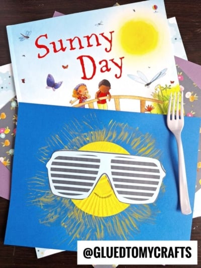 A children's book titled Sunny Day lies on colorful paper. Below it is a craft of a yellow sun with a smiling face and striped sunglasses made using fork painting crafts, placed on blue paper next to a pink plastic fork. The handle @GLUEDTOMYCRAFTS is at the bottom.