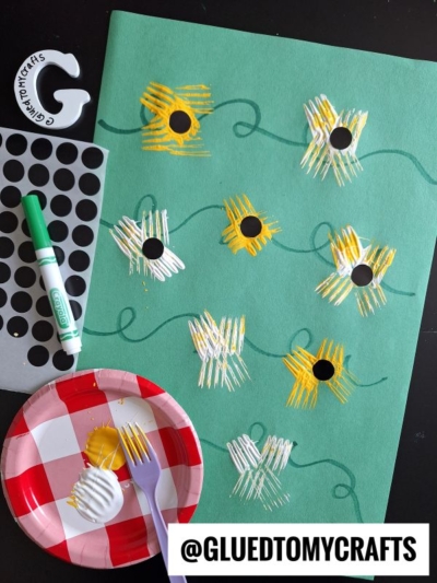 A green sheet features painted sunflowers created with yellow and white fork painting crafts and black centers. Nearby are a green marker, plastic fork, polka dot stickers, and a red paper plate. @GLUEDTOMYCRAFTS is at the bottom.