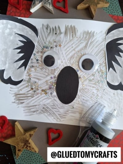 A handmade koala face crafted with white paint using fork painting crafts, black paper, large googly eyes, star glitter, and a plastic fork. Surrounded by gold and red stars and hearts. The handle @GLUEDTOMYCRAFTS is visible.