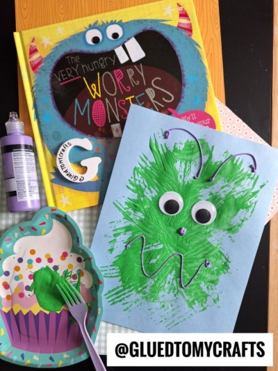 A children’s book titled “The Very Hungry Worry Monsters,” a purple paint bottle, a paper plate with green paint, a fork for fork painting crafts, and a blue paper with a green monster craft made from paint and googly eyes are shown.