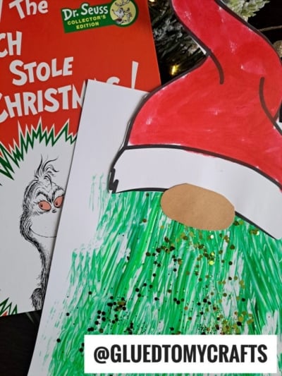 A Dr. Seuss How the Grinch Stole Christmas! book and a Grinch fork painting craft made with painted green lines, gold glitter, and a red Santa hat. The handle @GLUEDTOMYCRAFTS appears at the bottom.