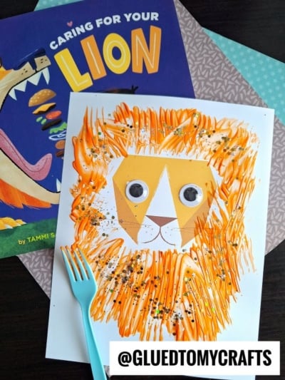 A handmade lion craft with an orange and yellow mane, created using fork painting crafts, googly eyes, and a paper face sits beside the book Caring for Your Lion. A plastic fork lies below the artwork on a dark surface.
