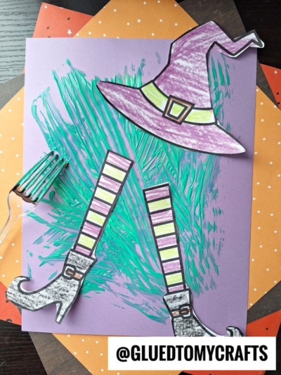 A Halloween craft with a purple paper background, fork painted green melted witch legs with striped stockings and pointed shoes, plus a purple witch hat. The cut-out legs and hat are arranged over splashes of green paint and orange polka-dotted paper.