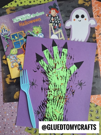 A Halloween-themed craft scene with a children's book, a ghost cutout, a fork painted monster hand on purple paper, black paper spiders, a blue plastic fork, and the @GLUEDTOMYCRAFTS handle.