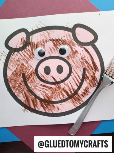 A pig face coloring sheet with brown paint brush strokes, googly eyes attached, and a plastic fork beside it—perfect for fun fork painting crafts. The image has the text “@GLUEDTOMYCRAFTS” at the bottom.