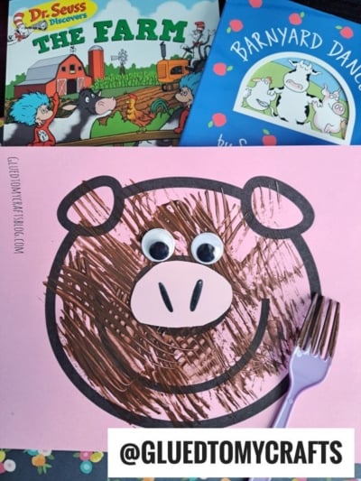A pink paper pig face is decorated with brown fork painting crafts strokes and two googly eyes. Behind it are children's books: The Farm and Barnyard Dance. A purple plastic fork and the @gluedtomycrafts logo are also visible.