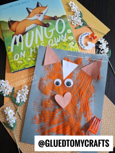 A children’s book titled Mungo on His Own sits next to a handmade fox craft with googly eyes and a heart-shaped nose, created using fork painting crafts and set on orange polka-dot paper with white flower decorations.