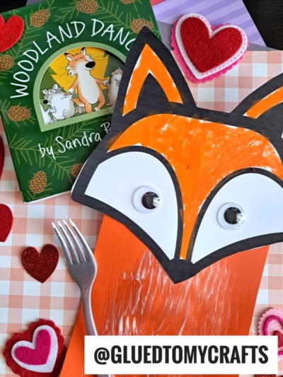 A children's fox craft made with paper, googly eyes, and fork painting crafts sits next to the book Woodland Dance! by Sandra Boynton. Felt heart decorations are spread on a pink checkered surface.
