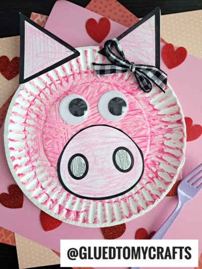 A paper plate pig craft with pink coloring, large googly eyes, a round snout, paper ears, and a black-and-white bow rests on heart-decorated paper alongside a purple fork—perfect for fun fork painting crafts! @GLUEDTOMYCRAFTS.