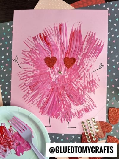 A pink monster with fork-painted fur, heart-shaped glitter eyes, and a small white nose is drawn on pink paper. Craft materials for fork painting crafts, including paint, a plastic fork, and heart shapes, are nearby. @GLUEDTOMYCRAFTS is labeled at the bottom.