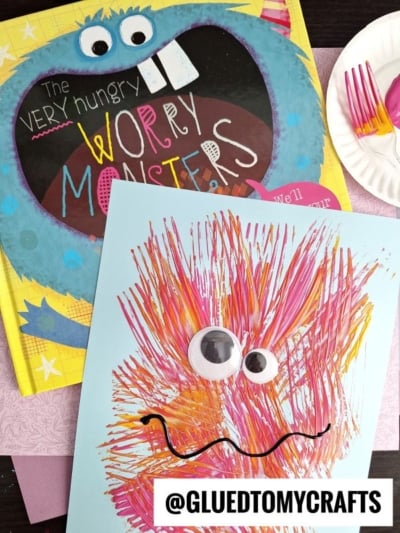 A colorful children’s book titled The Very Hungry Worry Monsters sits next to a blue paper monster, made with fork painting crafts—painted streaks, googly eyes, and a wavy mouth. Pink forks and a plate are in the background.