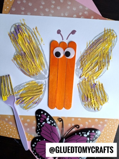 A butterfly craft made with orange popsicle sticks for the body, googly eyes, and wings fork painting crafts in yellow and purple; a pink plastic fork and a decorative butterfly are nearby.
