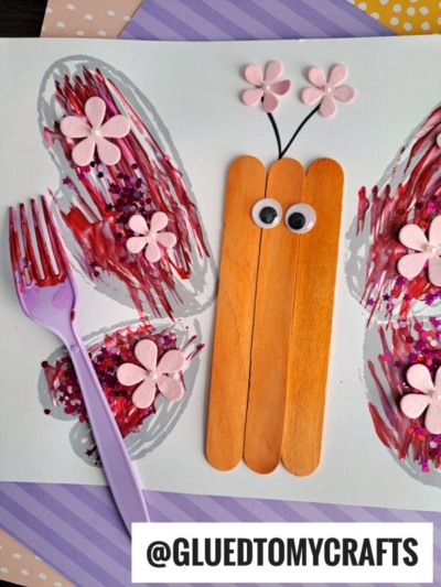 A butterfly craft made with popsicle sticks for the body, googly eyes, pink paper flowers for wings, and a purple fork painted using fork painting crafts technique, on a decorated background. @GLUEDTOMYCRAFTS is written at the bottom.
