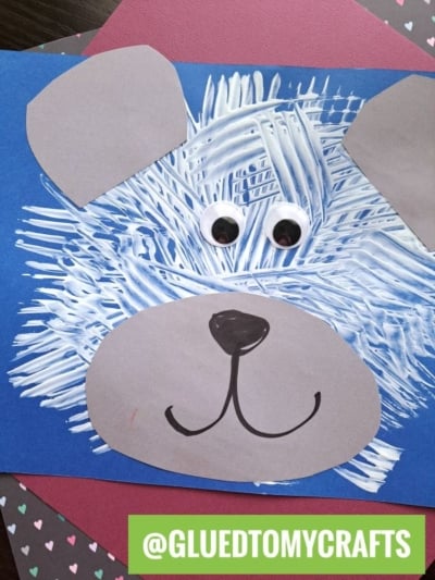 A children’s bear craft made with a blue paper background, fork painting crafts using white paint strokes for fur, gray paper ears and snout, googly eyes, and a drawn-on nose and mouth. A label reads @GLUEDTOMYCRAFTS.