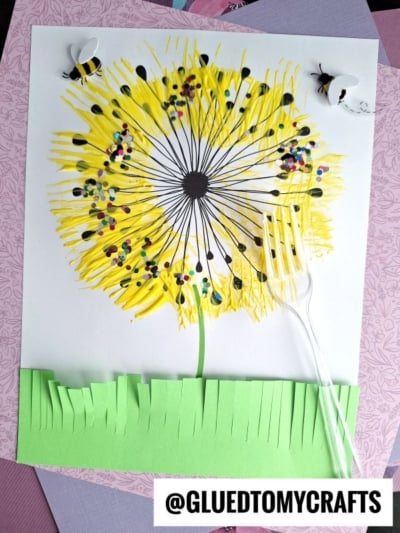 A colorful dandelion art project using fork painting crafts—yellow paint spread by a plastic fork, black dots, confetti, paper grass, and two paper bees. The fork handle is attached to the paper. @GLUEDTOMYCRAFTS is written at the bottom.