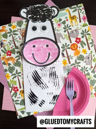 A cow craft made from white paper, black marker, googly eyes, and a pink rounded snout sits on colorful animal-printed paper. A pink plastic fork with black paint hints at fun fork painting crafts on the nearby pink plate. The text reads @GLUEDTOMYCRAFTS.