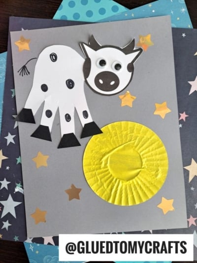 A handmade greeting card features a handprint cow with googly eyes and black spots, next to a yellow moon and gold stars on a gray background. @GLUEDTOMYCRAFTS is at the bottom.
