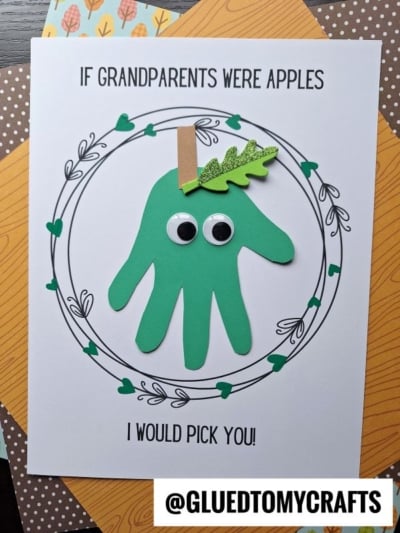 A craft card featuring a handprint apple made from green paper, complete with googly eyes and a paper leaf. Perfect for Grandparent’s Day, the card includes the sweet message: “If grandparents were apples, I would pick you!” The background is decorated with patterned paper layers for an extra special touch.