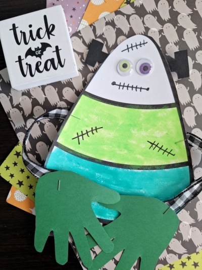 A colorful Halloween craft shaped like a Frankenstein monster, featuring googly eyes, handprint cut-outs for hands in a candy corn hug, and decorative stitching. It sits on ghost-patterned paper beside a sign that reads trick or treat.