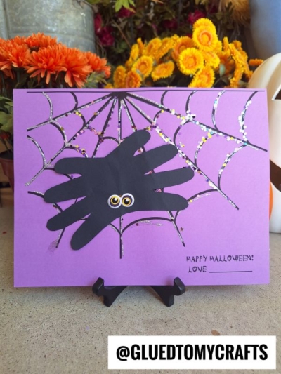 A purple card featuring a playful handprint spider with googly eyes sits on a web, with “Happy Halloween! Love ___” written in black. Orange flowers and festive Halloween decor create a cheerful background.
