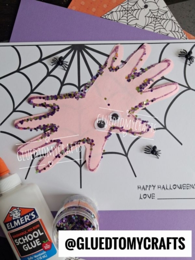 A handprint spider craft on spider web paper, decorated with glitter and toy spiders, alongside glue, purple paper, and a Happy Halloween message. @GLUEDTOMYCRAFTS tag is displayed at the bottom.
