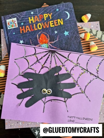 A children’s Halloween craft features a handprint spider with googly eyes on purple paper, decorated with a drawn spider web and colorful dots. In the background is a Happy Halloween book and scattered candy corn pieces.