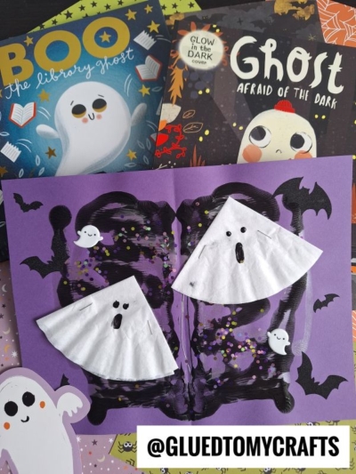 A purple Halloween craft with painted bats and two coffee filter ghosts made from white filters, surrounded by children's ghost-themed books and a cartoon ghost sticker. The handle @GLUETOMYCRAFTS is at the bottom.