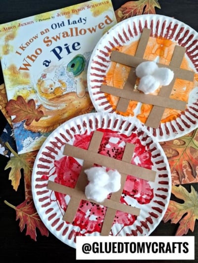 A children’s book titled I Know an Old Lady Who Swallowed a Pie sits next to crafty paint splat pie creations made from paper plates, cardboard strips as lattice, and cotton balls, displayed on a table with fake autumn leaves.