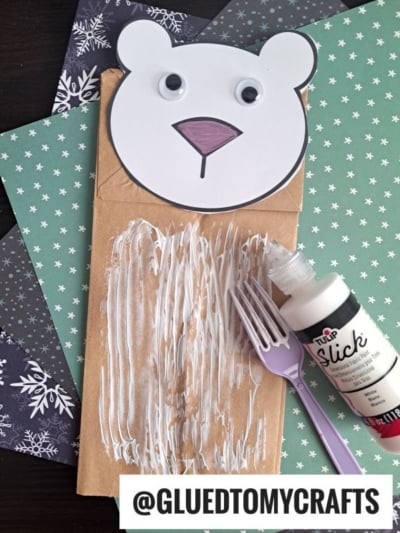 A paper bag with white paint spread on it, a purple plastic fork for fork painting crafts, a bottle of white fabric paint, and a polar bear face cutout sit on green and black snowflake-patterned paper. Text reads @GLUEDTOMYCRAFTS.
