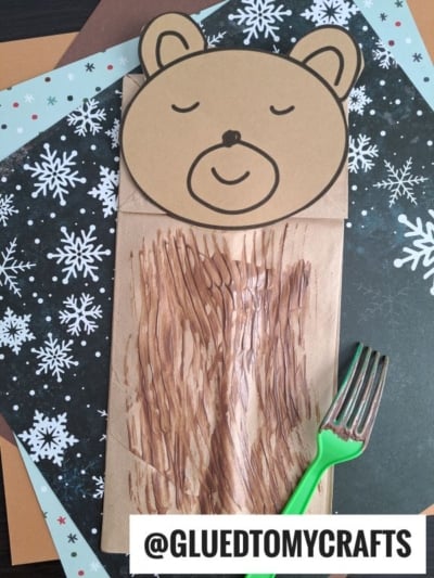 A paper bag bear craft featuring a round bear face and textured brown paint on the bag, created with fork painting crafts using a green fork. The background showcases snowflake patterns, with @GLUETOMYCRAFTS labeled at the bottom.