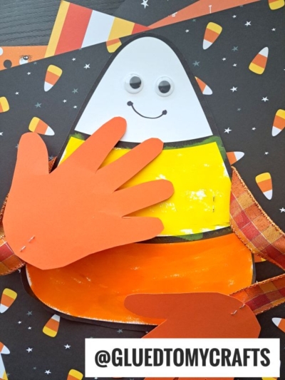 A candy corn craft with googly eyes, a smile, and orange paper handprints for arms gives the appearance of a cheerful candy corn hug, set on a black background with playful candy corn patterns.