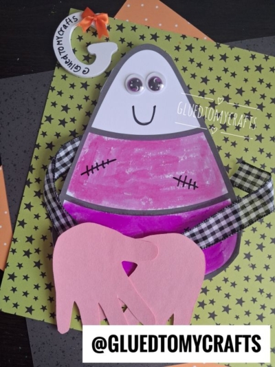 A pink and white paper candy corn craft with googly eyes, a smile, ribbon arms, and two pink handprint cutouts that create a sweet candy corn hug on a Halloween-themed background. Text @GLUEDTOMYCRAFTS appears on the image.